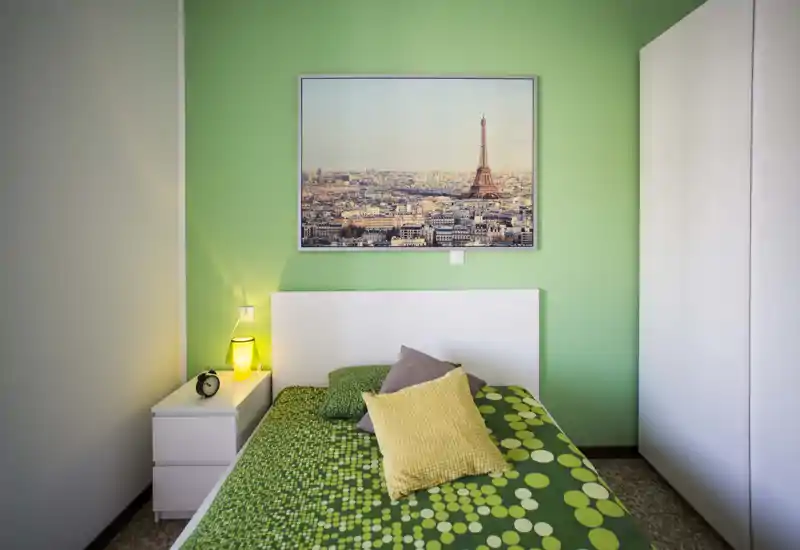 Chambre lumineuse et soignée avec mur vert, lit simple avec couette verte à motifs et coussins décoratifs, table de chevet avec lampe et réveil, et un tableau de la ville au-dessus de la tête de lit.