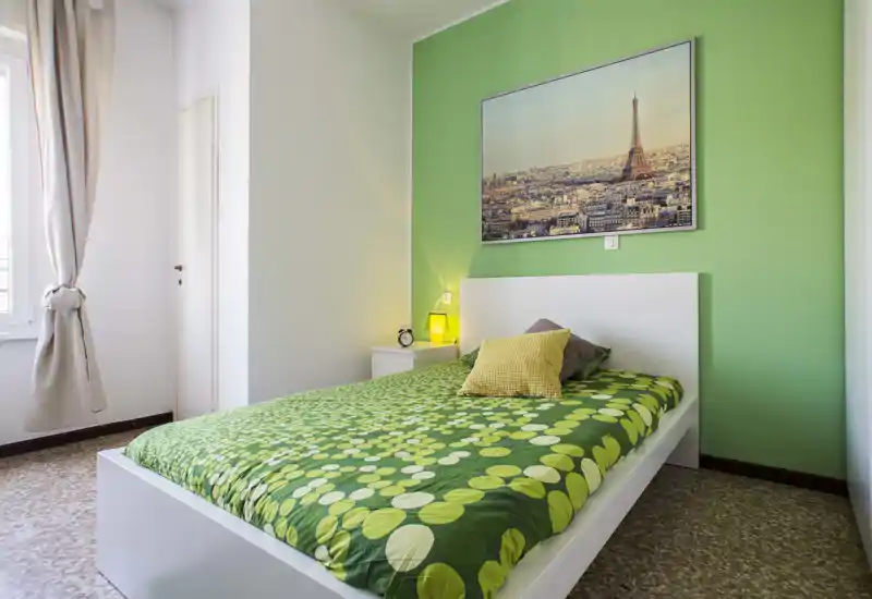 Chambre simple lumineuse et accueillante avec un mur vert, une couette verte à motifs, une table de chevet avec lampe et une photo de la ville au-dessus de la tête de lit.