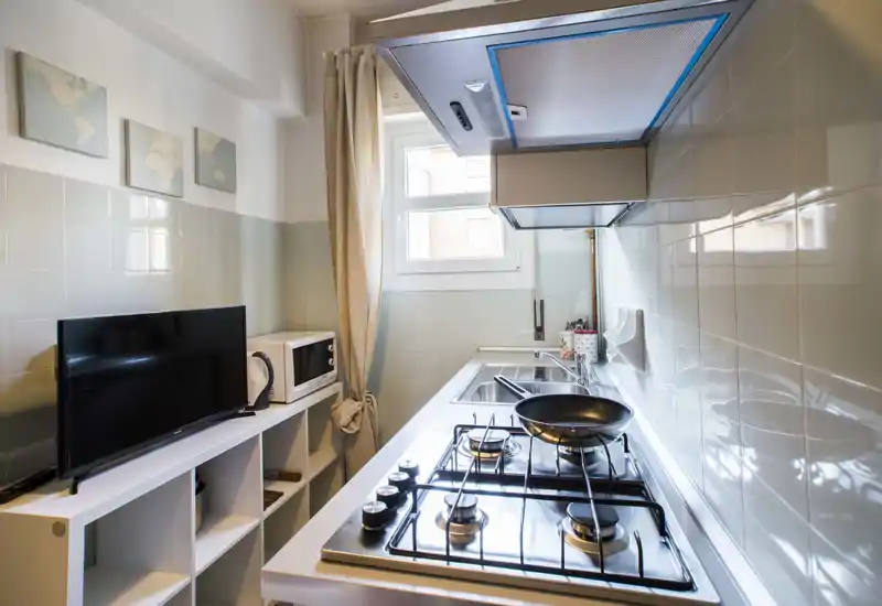 Compact modern kitchen with a four-burner gas stove, stainless sink, extractor hood and natural light from a window; tidy shelving with microwave and TV adds functional convenience.