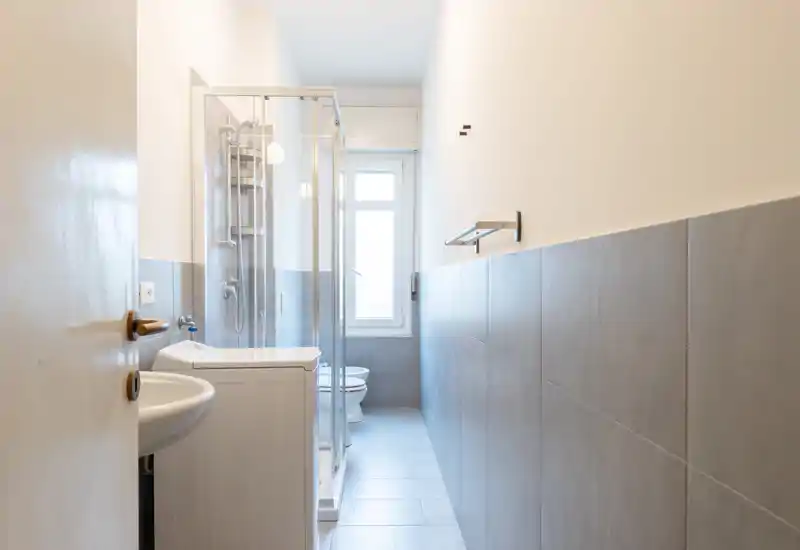 Bright, narrow bathroom with a glass shower enclosure, wall-mounted sink, washing machine and toilet near a window — clean modern tiling and neutral tones.