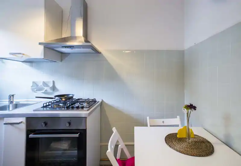 Compact modern kitchen with gas hob, built-in oven and sink. Light-colored tiled backsplash and a small dining table with decorative placemat and vase create a neat, welcoming space.