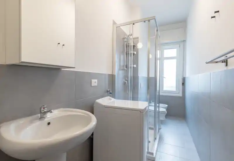Bright, modern bathroom with a glass shower enclosure, pedestal sink and toilet; neutral grey tiles and a window provide natural light.