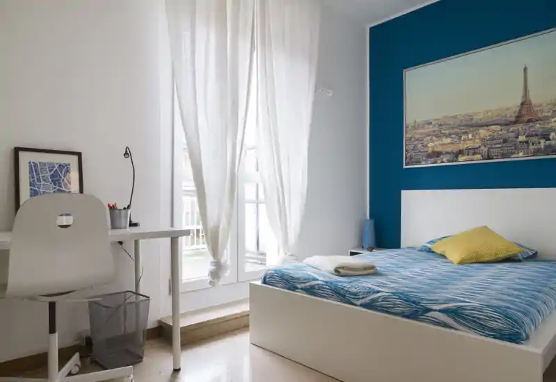 Dormitorio moderno y luminoso con cama blanca, ropa de cama azul con estampado y cojín amarillo. Gran ventana con cortinas translúcidas que aporta mucha luz natural; un escritorio pequeño y una silla crean una zona de trabajo.