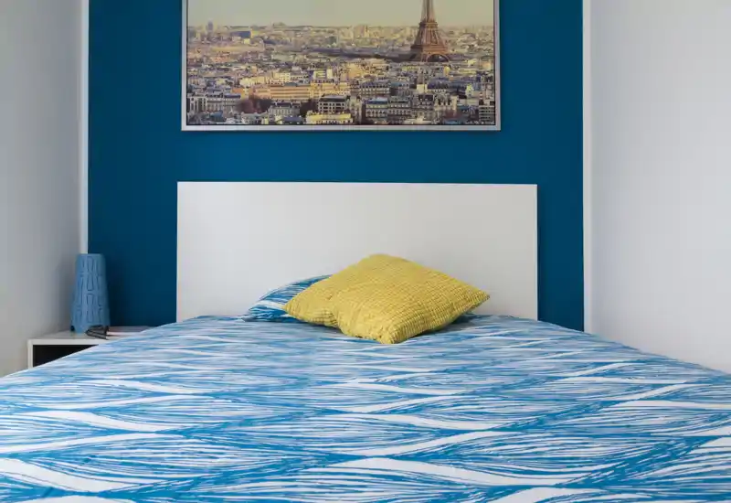 Dormitorio moderno y luminoso con una cama amplia vestida con ropa de cama azul estampada, cojín amarillo y un cuadro de la ciudad sobre la cabecera; líneas limpias y contraste de color que crean un ambiente fresco y acogedor.