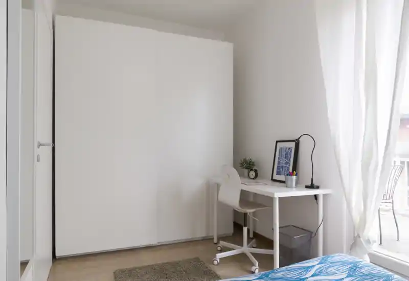 Dormitorio luminoso y minimalista con armario blanco, un pequeño escritorio y cama parcialmente visible con ropa de cama azul; espacio limpio, moderno y bien iluminado, ideal para una persona.