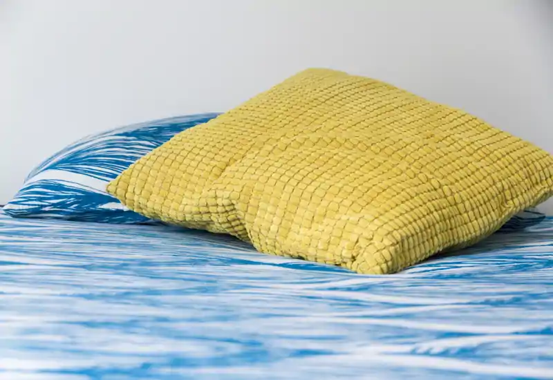 Plano cercano de una cama bien hecha con sábanas azules estampadas y dos cojines amarillos texturizados, mostrando el detalle de las telas y una pared neutra.