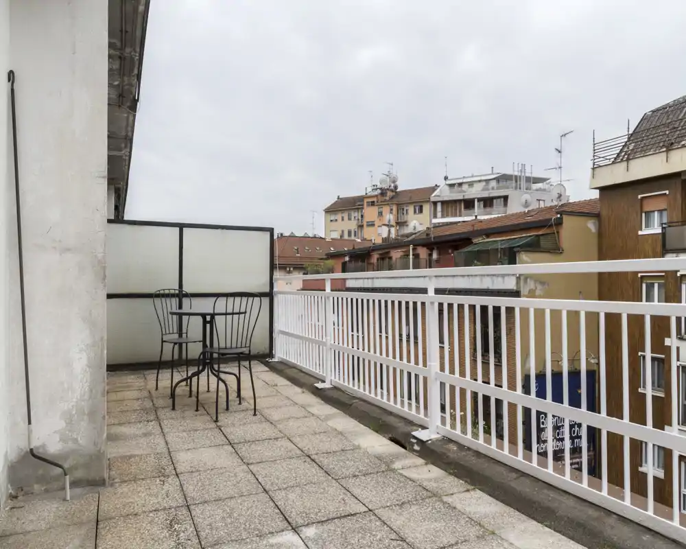 Balcone ampio con ringhiera metallica e piccolo tavolo da bistrò con sedie, vista sui tetti della città — perfetto per il caffè mattutino.