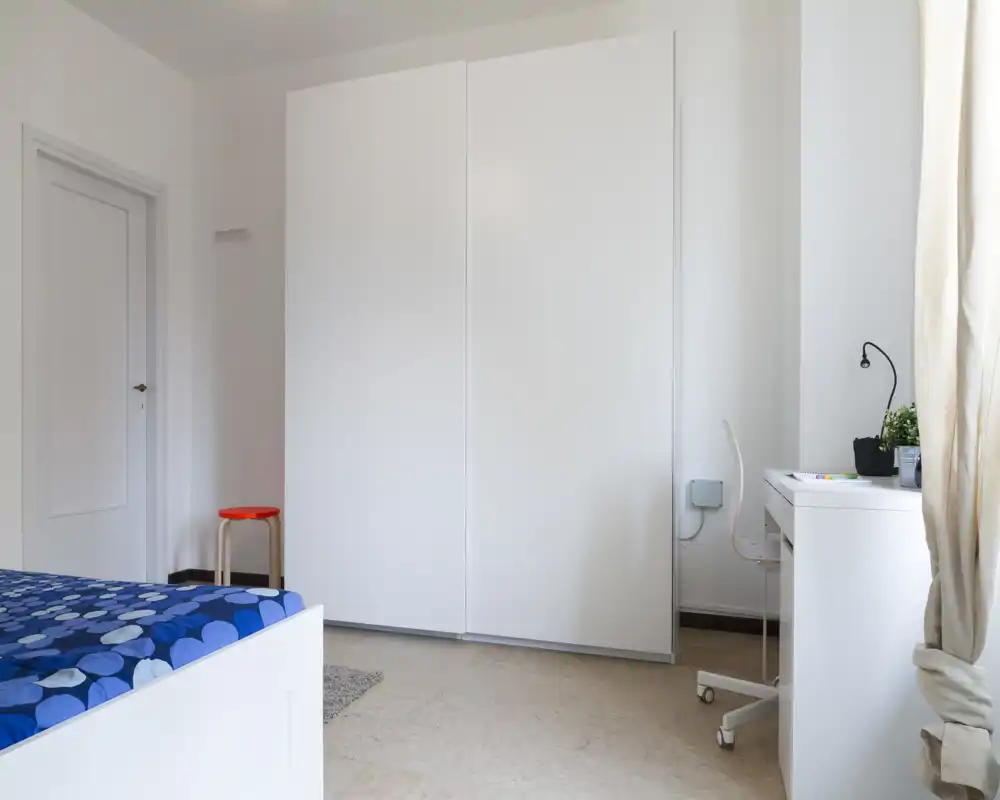 Camera da letto minimalista con grande armadio bianco, letto singolo con copriletto blu a motivi e una piccola scrivania vicino alla finestra; luminosa e ordinata.