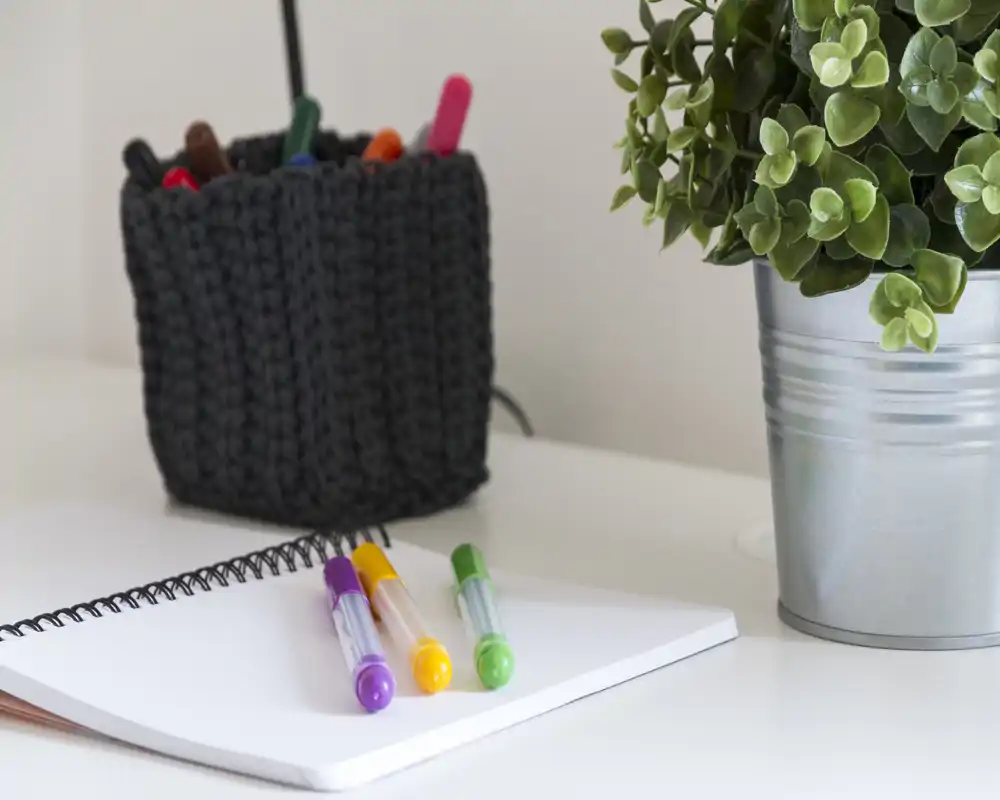 Primo piano di una superficie d'ingresso o di lavoro con un taccuino, pennarelli colorati e una pianta in vaso — una vignetta semplice e luminosa perfetta per mostrare un angolo scrivania o un tavolino d'ingresso.