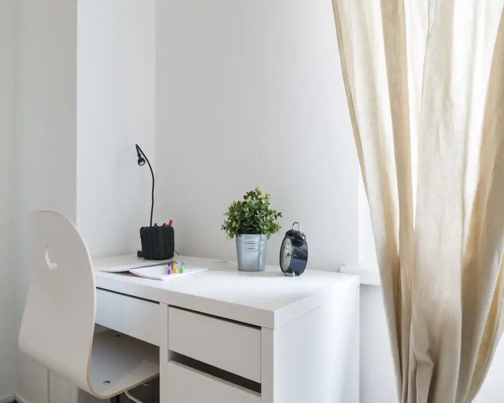 Angolo studio luminoso e minimale con scrivania bianca, sedia, pianta in vaso e sveglia accanto a una finestra con tende beige chiare.