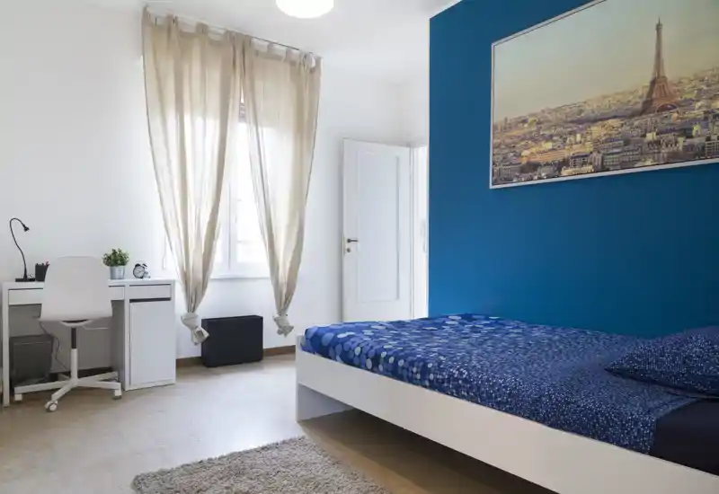Camera da letto moderna e luminosa con parete blu e letto singolo ben rifatto; una scrivania vicino alla finestra offre un comodo spazio di lavoro con luce naturale.