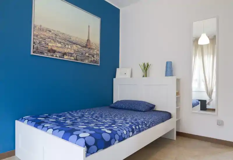 Camera da letto singola luminosa e ordinata con letto dalla struttura bianca su una parete accentata blu, biancheria a motivi blu, quadretto e uno specchio che riflette luce naturale.