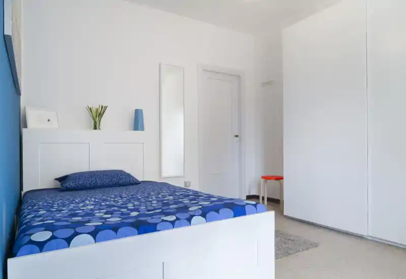 Camera da letto luminosa e moderna con letto in cornice bianca e biancheria blu a motivo, uno specchio alto e arredi minimalisti — pulita e ariosa.
