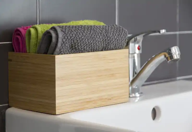 Close-up of a bathroom sink area showing a chrome faucet, tiled backsplash and a wooden box holding neatly folded towels in gray, green and pink.