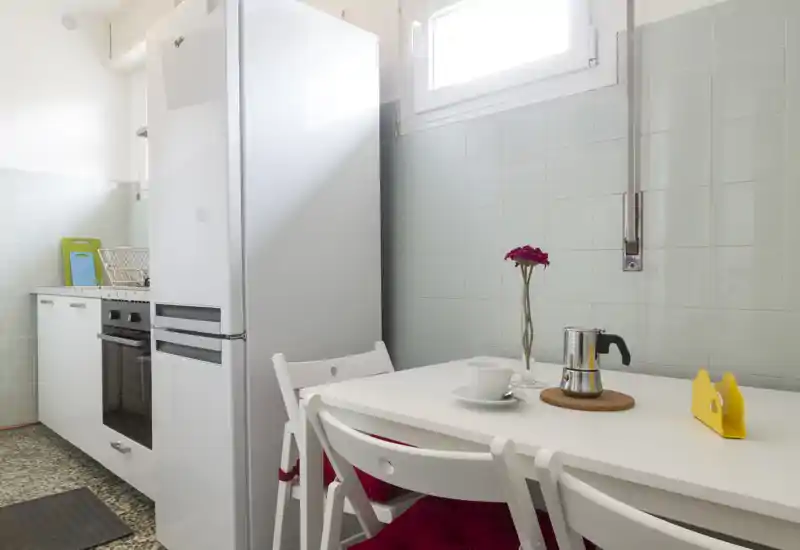 Compact, well-lit kitchen corner with white cabinets, refrigerator and a small dining table set for one. Clean, minimal styling with a fresh vase and bright cushions.