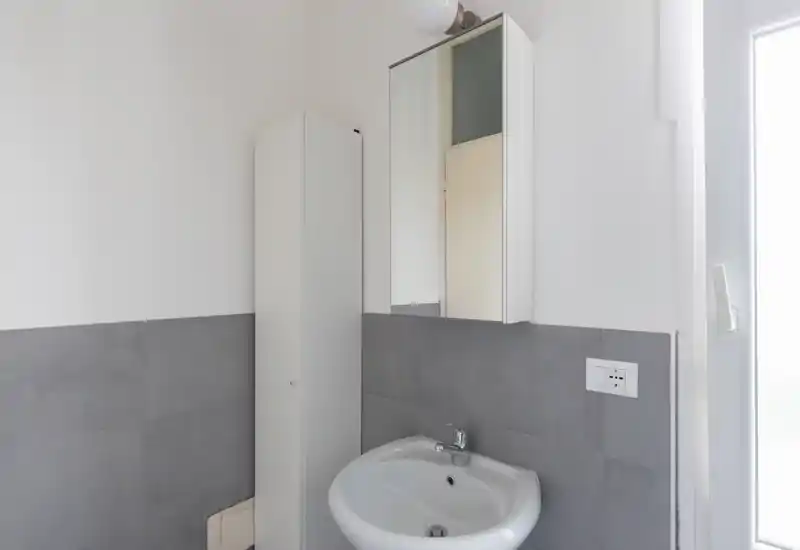 Small modern bathroom with a wall-mounted sink, mirrored cabinet and tall storage unit; neutral gray tiles and bright natural light from a window.