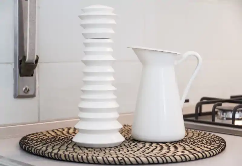 Close-up of a clean kitchen countertop with decorative white jug and vase beside a gas hob, staged on a woven placemat.