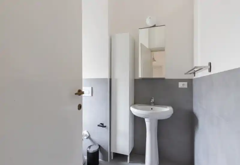 Small, clean bathroom with a pedestal sink, mirrored cabinet and compact storage; neutral grey tiles and white walls provide a minimalist, well-lit space.