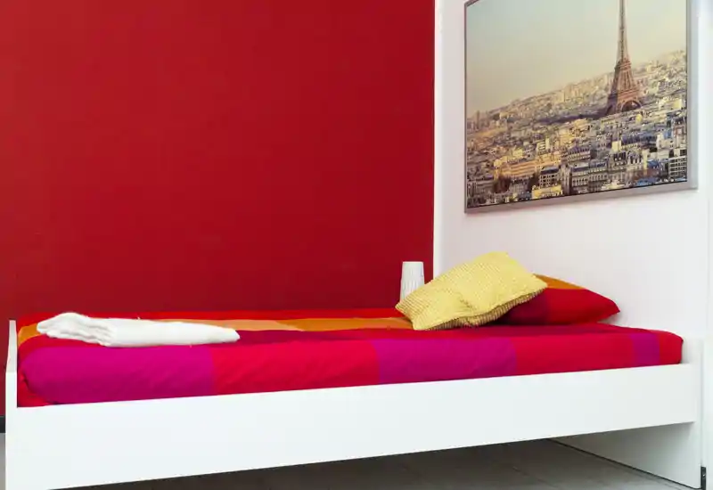Camera da letto singola luminosa e ordinata con letto bianco, biancheria colorata e una stampa incorniciata di una città sulla parete. Parete d'accento rossa e arredo minimal ideale per un affitto compatto.
