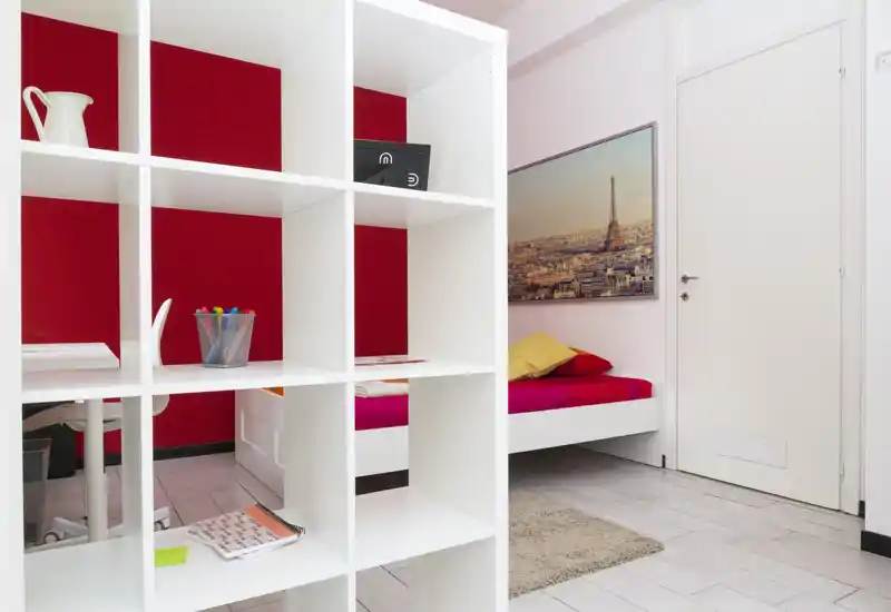 Camera da letto moderna e luminosa con una parete rossa, letto singolo con biancheria vivace e cuscini decorativi, e una libreria bianca a vista che suddivide parzialmente lo spazio. Linee pulite, un tappeto morbido e un quadro rendono l'ambiente accogliente e ideale per affitti brevi.