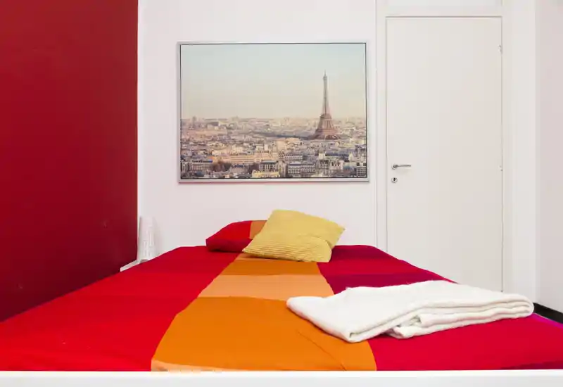 Camera da letto luminosa e ben allestita con letto matrimoniale dai colori vivaci, cuscini decorativi e una stampa di Parigi sopra la testiera. Linee pulite e biancheria rossa-arancione creano un ambiente moderno e accogliente, perfetto per annunci.