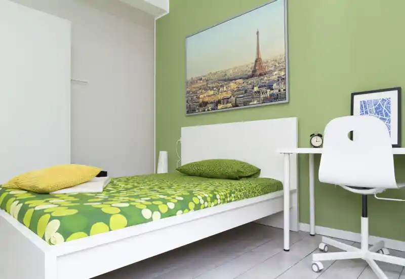 Chambre simple moderne et lumineuse avec lit aux accents verts, cadre blanc, bureau et chaise — style minimaliste propre et tableau de ville au-dessus de la tête de lit.