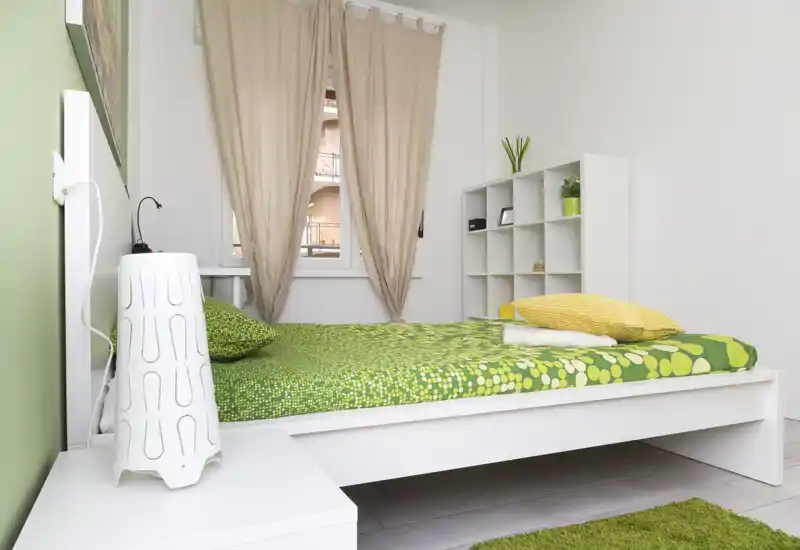 Chambre moderne et lumineuse avec lit blanc à literie verte à motifs, coussin jaune, petite lampe de chevet et étagère modulable — lumière naturelle et décoration minimaliste.