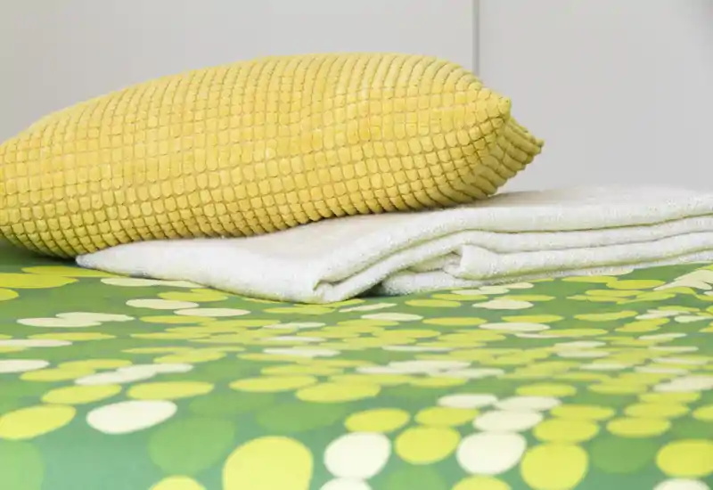 Gros plan sur des serviettes pliées et un coussin jaune texturé posés sur une couette verte à motifs — détail soigné de la chambre montrant du linge frais et des textiles colorés.