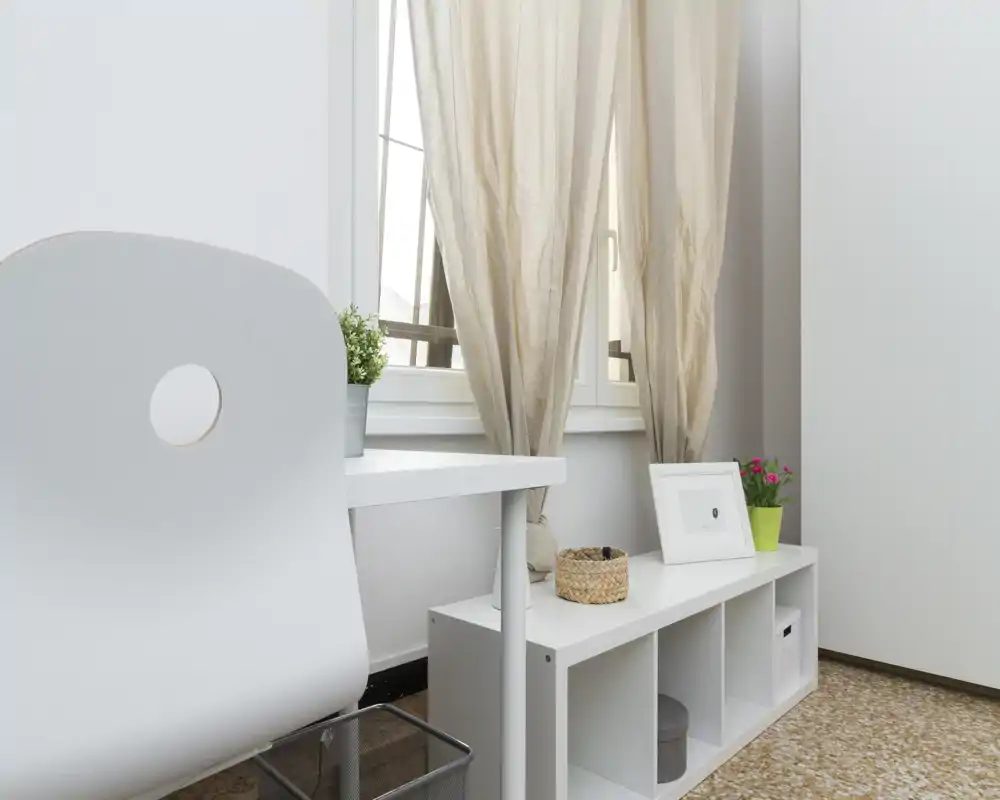 Angolo living minimale e luminoso con scrivania bianca, sedia moderna e mensola contenitore sotto una finestra che diffonde luce naturale.