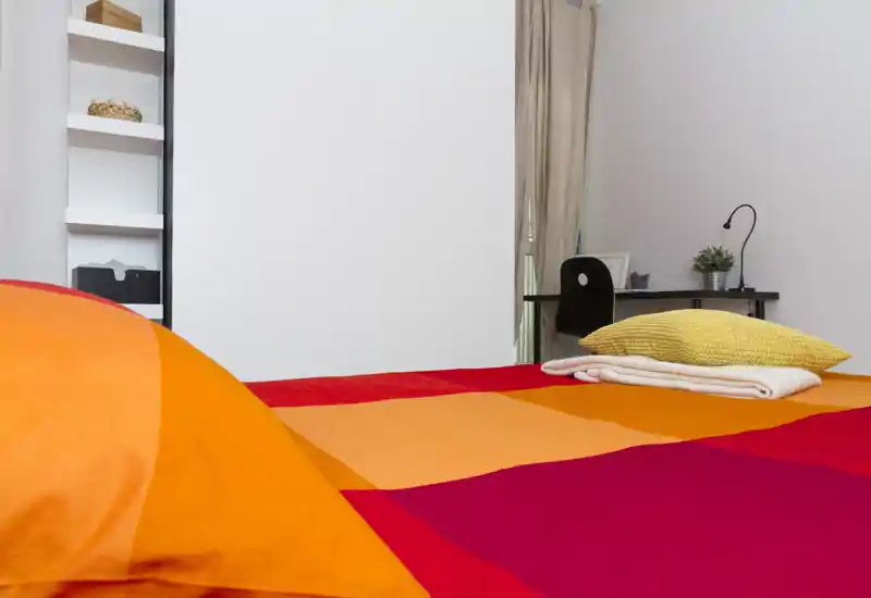 Chambre moderne et lumineuse avec une couette colorée, du linge plié et un petit bureau près de la fenêtre — propre, accueillante et bien éclairée.