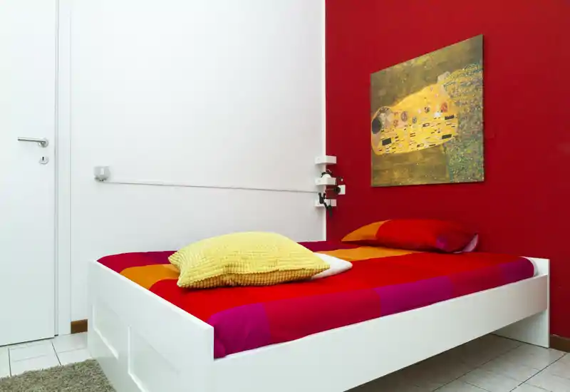 Chambre moderne et lumineuse avec un lit simple blanc habillé de linge rouge-orangée et un coussin jaune. Sol carrelé propre et mur d'accent rouge avec tableau, espace soigné et accueillant pour une annonce locative.