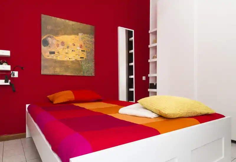 Chambre moderne et lumineuse avec un lit blanc habillé d'une couette rouge-orangée intense, des coussins moelleux et une œuvre décorative sur un mur rouge profond. La composition met l'accent sur le lit et les textiles chaleureux.