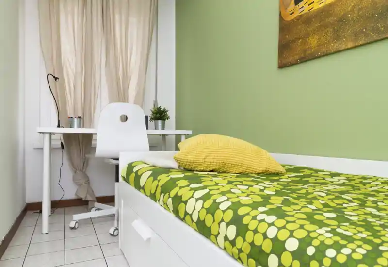Dormitorio compacto y luminoso con cama individual verde estampada, cojín amarillo y pequeño escritorio blanco junto a la ventana — decoración sencilla y fresca, ideal para estudiantes o estancias cortas.