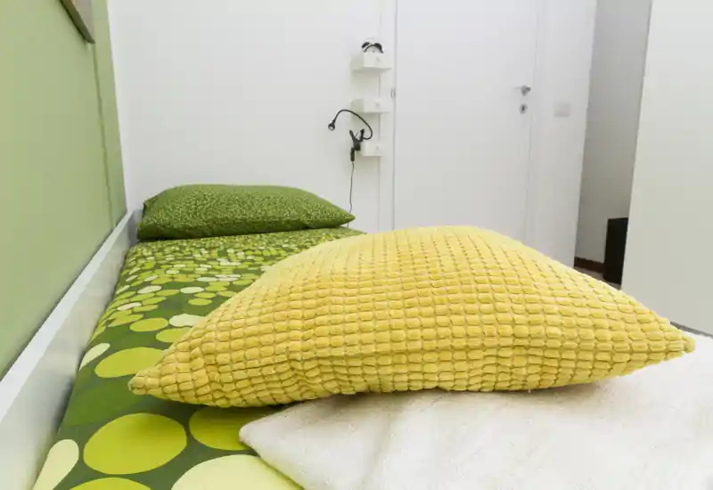 Dormitorio compacto y luminoso con una cama individual con sábanas verdes estampadas y un cojín amarillo texturado. Paredes blancas y estanterías minimalistas que ofrecen un aspecto moderno y fresco, perfecto para estancias cortas.