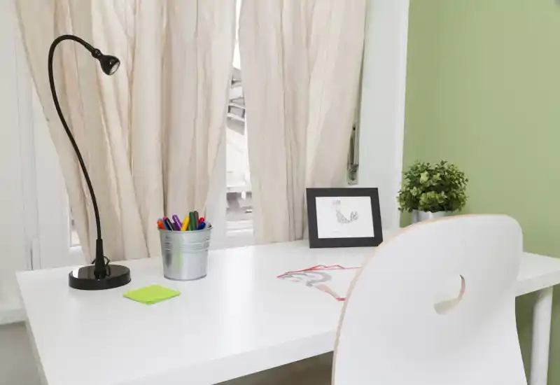 Espacio de trabajo luminoso y ordenado con escritorio blanco, lámpara, bolígrafos de colores en un vaso metálico, cuadro enmarcado y una pequeña planta junto a una ventana con cortinas claras.