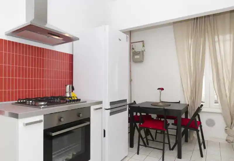 Cucina compatta e moderna con paraschizzi in piastrelle rosse, piano cottura a gas in acciaio e forno, frigorifero bianco e piccolo tavolo da pranzo con sedie con cuscini rossi — luminosa e pratica per la vita in città.