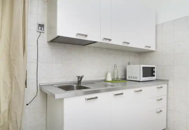Angolo cottura compatto e moderno con mobili bianchi, piano e lavello in acciaio inox e microonde — soluzione pulita e pratica per soggiorni brevi.