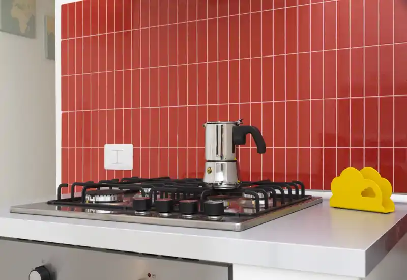 Dettaglio di cucina moderna compatta con piano cottura a gas e moka davanti a una piastrella rossa lucida. Piano pulito e stile luminoso e contemporaneo, perfetto per un annuncio di affitto.