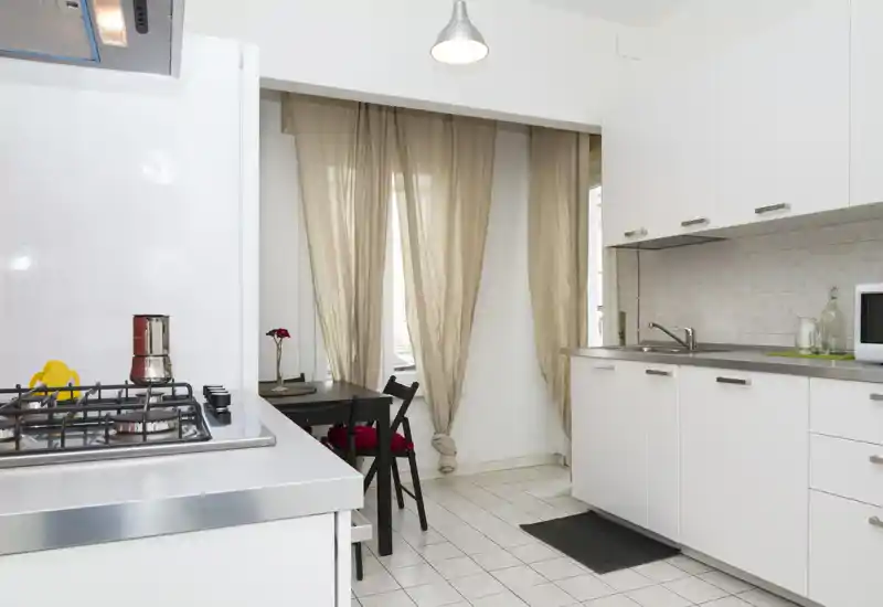 Cucina moderna luminosa con pensili bianchi, piano in acciaio inox e piccolo tavolo da pranzo vicino alla finestra; ordinata e ben illuminata.