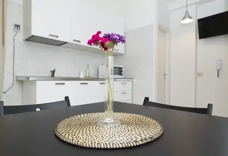 Cucina compatta e luminosa con pensili bianchi, piano in acciaio e un piccolo tavolo da pranzo decorato con un vaso di fiori — pulita, moderna e accogliente.