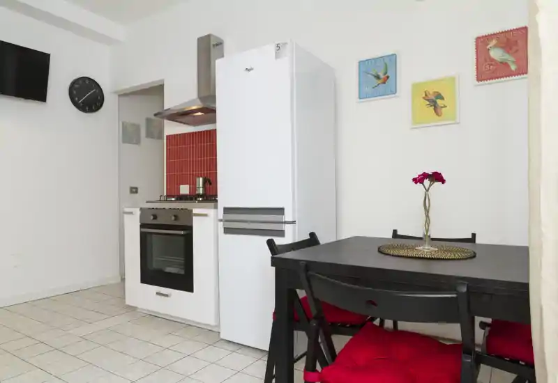 Angolo cottura moderno e compatto con piano a gas, forno, frigorifero alto e tavolo scuro con sedie con cuscini rossi; pareti bianche luminose e quadretti colorati rendono l'ambiente accogliente.
