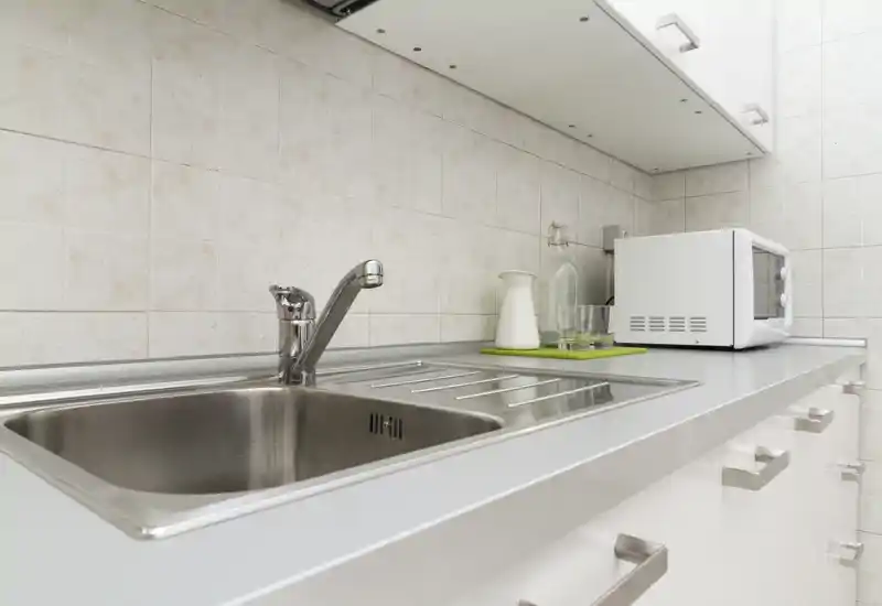 Bancone cucina compatto e luminoso con lavello in acciaio inox, miscelatore moderno, piastrelle a parete e microonde sul piano di lavoro.