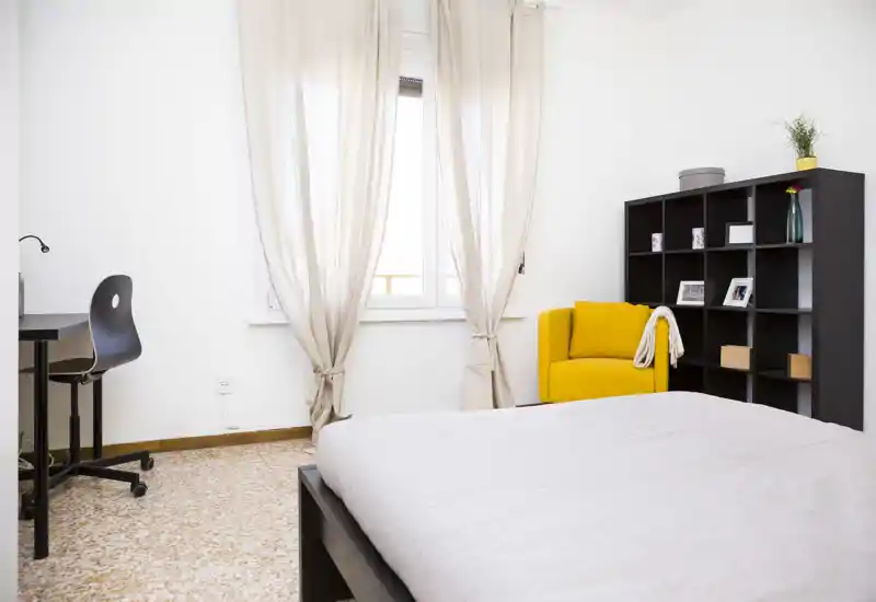 Bright, modern bedroom with a large bed in the foreground, a desk by the window and a bold yellow armchair beside an organized shelving unit.