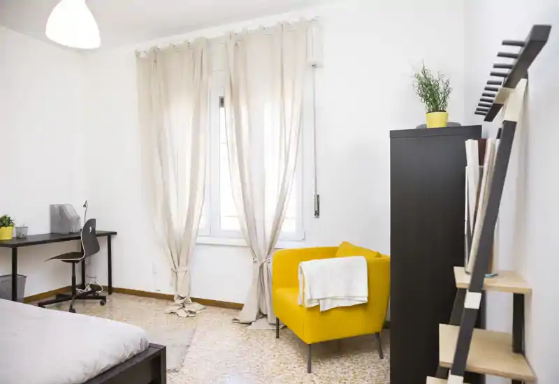 Bright, modern bedroom with a comfortable bed, a sunny window dressed with light curtains and a cheerful yellow armchair creating a cozy reading corner.