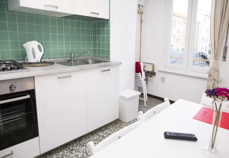 Compact, modern kitchen with white cabinets, stainless sink, green tiled backsplash and a small dining table by a bright window — well organized and ready for use.