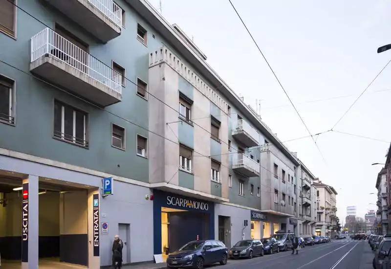 Street-level exterior showing a multi-story building with ground-floor retail (ScarpaMondo), parked cars and balconies — a clear urban storefront scene.