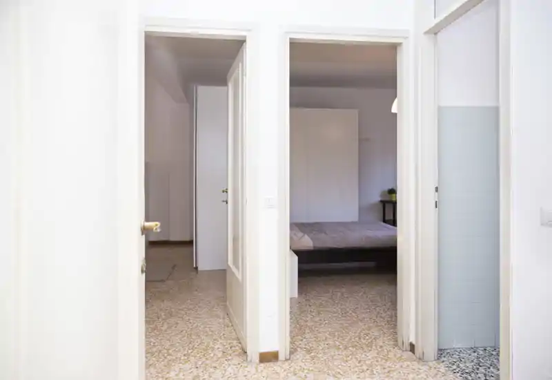 View through a corridor into a bedroom with a double bed visible through the doorway; neutral linens, a bedside table and bright, clean walls create a calm, minimal space.