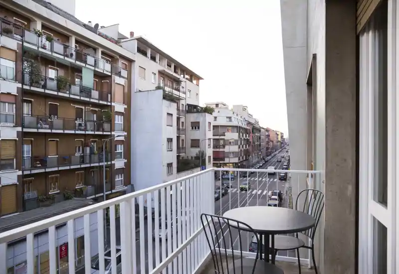 Small urban balcony with a round metal table and two chairs, overlooking a busy street and neighboring apartment buildings.
