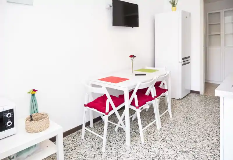 Small bright dining area with a white table and four chairs featuring red cushions, a wall-mounted TV and a tall fridge nearby — a compact, functional space ideal for casual meals.