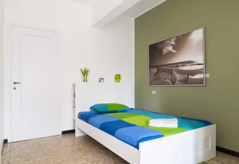Camera da letto singola luminosa e ordinata con struttura del letto bianca, biancheria colorata blu-verde, quadro a parete e arredo minimale — ideale per una persona o soggiorni brevi.